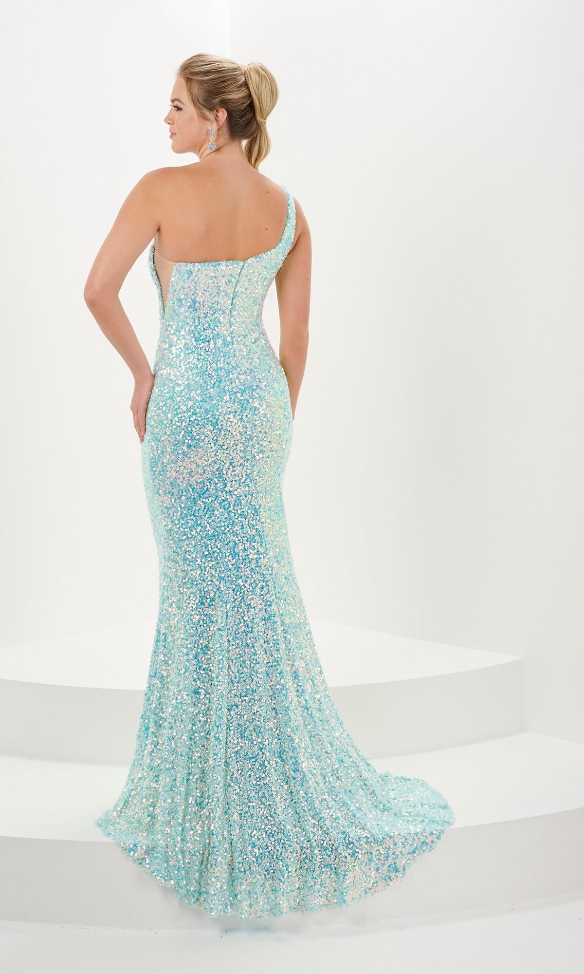 A woman stands on a white platform, shown from the back, wearing the Tiffany One-Shoulder Long Sequin Prom Dress 16114 in light blue with a fitted silhouette and floor-length design.